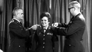 Two military officers in uniform pin new rank insignia on a smiling woman in military attire, who stands between them in front of a curtain during a formal ceremony.