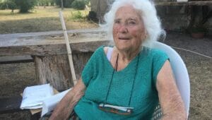 An elderly woman with white hair sits outside on a white chair, wearing a green top and glasses, near a wooden table and garden.