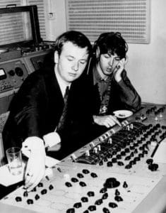 Two men sit at a large audio mixing console. One, in a suit, adjusts controls while Paul McCartney, wearing headphones, listens and observes. Recording equipment and a glass of water are nearby. The scene appears to be a 1960s era recording studio.
