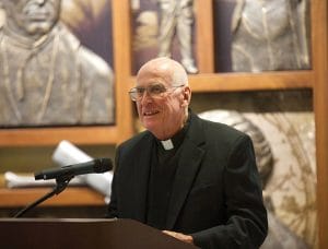 An elderly man in glasses and a black clerical suit with a white collar smiles, speaking at a lectern. Art is seen behind him.