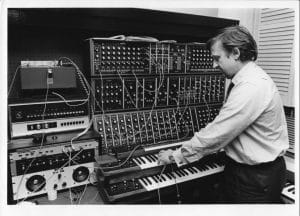 A man operating a large modular synthesizer with numerous knobs, cables, and switches in a studio. He is adjusting controls while standing next to an electronic keyboard and audio equipment.