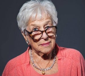 Elderly woman with short white hair wearing tortoiseshell glasses, a coral top, pearl necklace, and earrings, smiling slightly against a dark gray background.