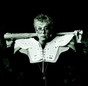 An older woman wearing glasses and football shoulder pads holds a baseball bat across her shoulders, looking directly at the camera with a serious expression. The image has a dark, high-contrast, monochrome tone.