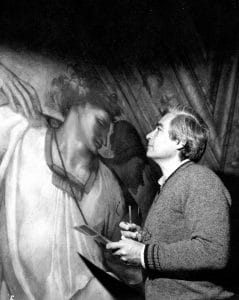 A man with cleaning tools examines a large mural of a classical figure in flowing robes. The black-and-white scene suggests restoration.