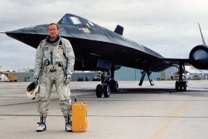 A pilot in a silver flight suit stands on a runway with a helmet and yellow case, in front of a sleek, black SR-71 Blackbird aircraft on an overcast day.