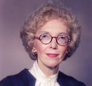 A woman with short, curly blonde hair, round glasses, and gold earrings poses in a dark robe and white blouse with a bow.
