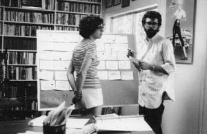 A woman and a man stand in front of a large corkboard covered with note cards in a room lined with bookshelves. The man holds a notepad and pen while they discuss the arrangement.