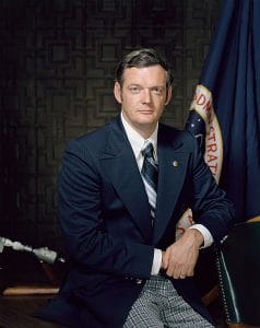 A man in a navy suit and striped tie sits before a dark backdrop with a NASA flag, looking at the camera with a neutral expression for a formal portrait.
