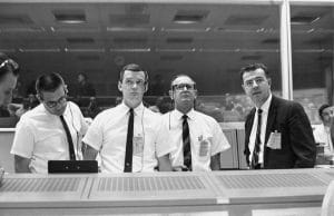 Four men in white shirts and ties stand at 1960s NASA mission control panels, looking off-camera; glass windows and others behind them.