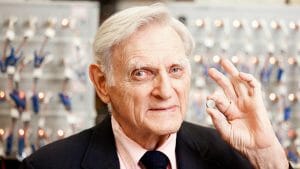 An elderly man in a suit smiles as he holds up a small round object, with blurred electronics and twinkling lights in the background.