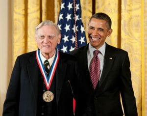 An older man wearing a medal stands beside a smiling President Obama, with an American flag and gold curtains behind them.