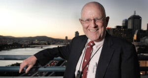 A smiling bald man in a suit and tie stands on a balcony at sunset, city buildings, river, and distant mountains behind him.
