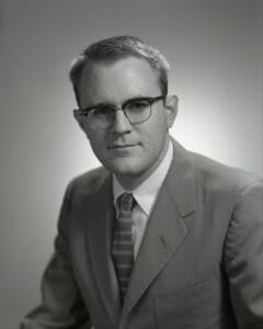 A black-and-white portrait of a man with glasses, short hair, in a suit and striped tie, facing the camera against a plain backdrop.