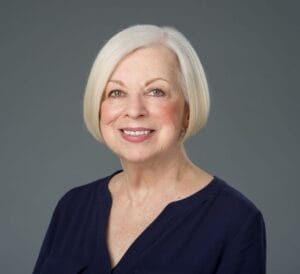 Smiling older woman with short, straight white hair and a dark blue blouse, standing in front of a simple gray backdrop.