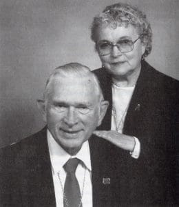 An older man in a suit and tie sits while an older woman stands behind him, hand on shoulder; both smiling in glasses, plain backdrop.