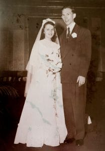 A vintage photo of a smiling bride in white with bouquet beside groom in dark suit, posing for a formal wedding portrait.