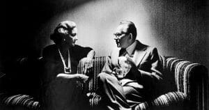 A woman in a dress and pearls and a man in a suit and glasses talk intently on a striped couch under dim lighting. The photo is black and white.