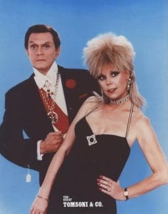 A man in a gaudy outfit including a tuxedo with a red sash, large gold necklaces, a red rose in his lapel poses with a woman in a black dress with a bold necklace against a blue background. The image reads “THE GREAT TOMSONI & CO.” at the bottom.
