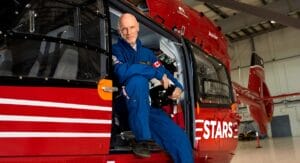 A man in a blue flight suit sits in the open doorway of a red STARS air ambulance helicopter in a hangar, smiling.