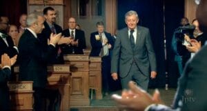 An older man in a suit walks down an aisle in a formal chamber as people on both sides applaud; nameplates and cameras visible.