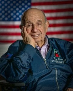 An elderly man with a gentle smile rests his head on his hand, wearing a blue jacket, seated before an American flag.