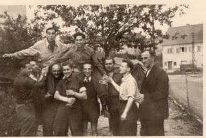 Nine people pose outdoors; seven stand while two are lifted on shoulders, smiling joyfully. Trees and houses in black-and-white 1940s-era backdrop.