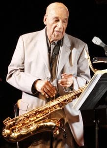 An older man in a light suit holding a brass saxophone in front of a microphone, preparing for or finishing a performance.