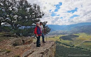 Two hikers stand on a rocky cliff edge with backpacks and trekking poles, overlooking a valley dotted with trees, fields, and distant mountains beneath a partly cloudy sky.