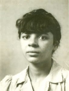 A black and white portrait of a young woman with dark hair and bangs, wearing a striped collared shirt, looking directly at the camera with a neutral expression.