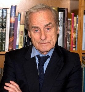An older man in a suit and tie stands with arms crossed before a bookshelf filled with architecture and design books.