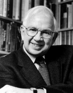 An older man with white hair and glasses smiles, wearing a dark suit and patterned tie, seated before shelves of books.