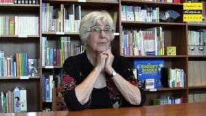 An elderly woman with gray hair and glasses sits at a wooden table by bookshelves. Her hands are clasped under her chin. A sign reads Charles M. Schulz Museum.