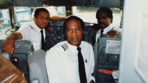 Three uniformed airline pilots sit in a plane cockpit, facing the camera with controls visible. They appear focused and professional.