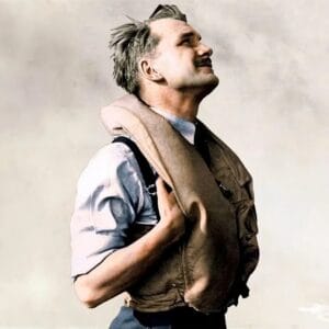 A man in profile with neat hair and a mustache looks upward, smiling, in a light brown life vest over a blue shirt on a plain background.