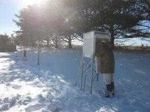 A person in winter clothing checks a weather instrument shelter in a snowy field, with trees in the background and bright sunlight overhead.