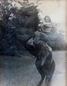 Black and white photo of a girl on a rearing horse outdoors, surrounded by trees and grass. The rider, who has long hair, is wearing a patterned shirt.