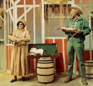 An older woman in a floral dress and bonnet and an older man in green jeans, matching jacket, and hat stand on a rustic stage, each holding a fiddle; between them sits a barrel and an open instrument case.