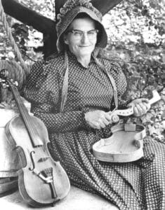 An older woman in a patterned dress and bonnet sits outdoors, smiling as she assembles a fiddle. Next to her on the bench lies a finished fiddle, with leafy branches surrounding them.