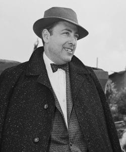A man wearing a wide-brimmed hat, bow tie, and a dark, textured overcoat smiles while standing outdoors. The background is slightly blurred, with some buildings and trees visible.