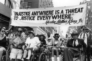A diverse group, many in wheelchairs, marches down a city street with a banner quoting MLK Jr.: 'Injustice anywhere is a threat to justice everywhere.'