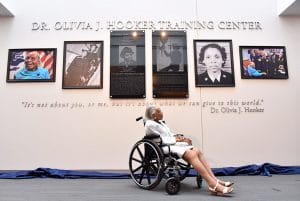 An elderly woman in a wheelchair looks up at a wall with photos and plaques honoring herself at the Dr. Olivia J. Hooker Training Center. A quote and six framed displays are mounted above her.