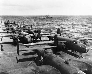 World War II bombers are lined up on the deck of a naval aircraft carrier at sea, with ocean waves and a distant ship visible in the background.