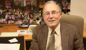 An older man in glasses, a brown blazer, and tie sits at an office desk with photos on the wall and papers spread before him.