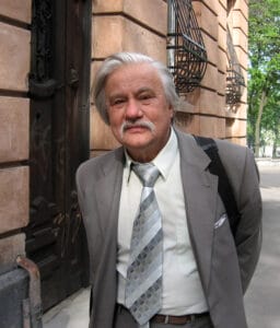 An older man in a gray suit and patterned tie stands outside a stone building with large windows; trees and a sidewalk are behind him.