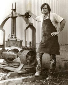 A man in an apron and work clothes stands outdoors by a vintage machine, hand resting on it, corrugated wall behind.