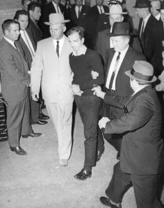 A group of men escort a handcuffed man in a dark sweater. Another man in a suit and hat points a handgun at the handcuffed man as a crowd looks on. The scene appears tense and chaotic.