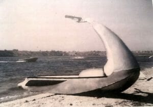 A black-and-white photo shows a whale-shaped single-person boat resting on sandy shore, with water and another boat in the background.
