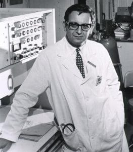 A man in a white lab coat and glasses stands beside laboratory equipment, with a clipboard and pen in his pocket. He appears to be in a scientific or medical laboratory.
