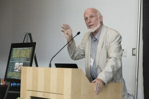 An older man with a white beard and light suit speaks at a lectern, gesturing. A display board with text and images is behind him.