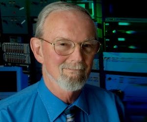 An older man with glasses, a short white beard, and a mustache wears a blue shirt and striped tie, standing in front of a background of electronic equipment and computer servers.